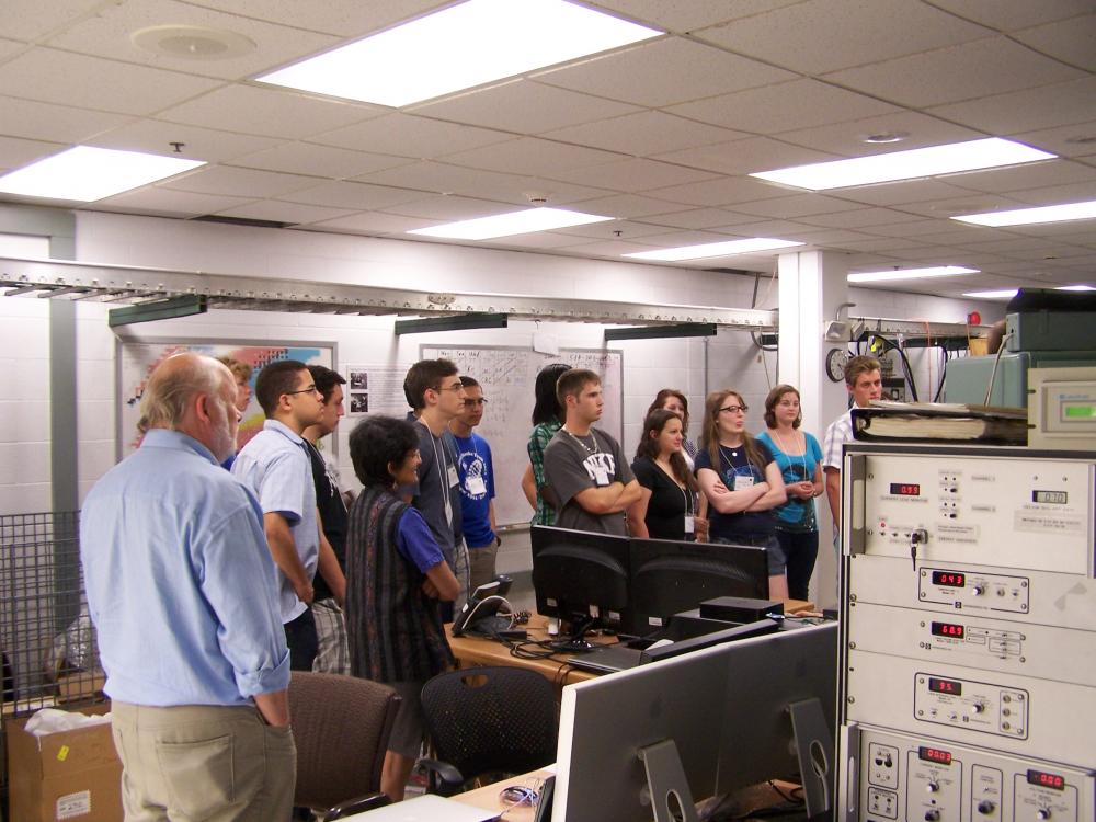 A group of students and chaperones looking toward an off-frame subject, obscured behind machinery, in a classroom computer lab